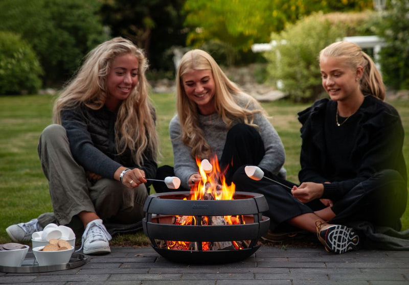 3 glade piger siddende rundt om bålfad og rister skumfiduser
