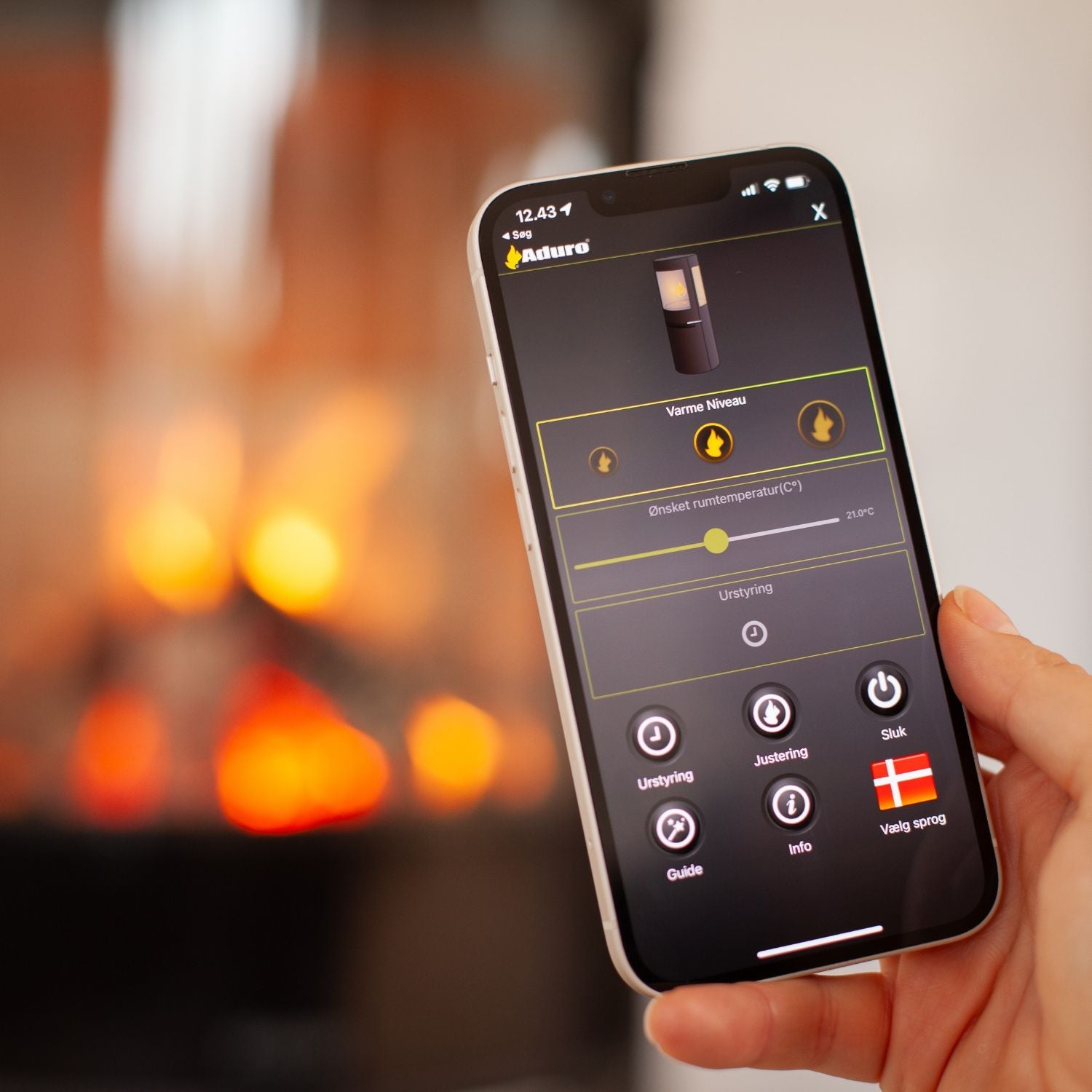 Hånd holder smartphone med hybrid app, der styrer temperaturen på en Aduro hybridovn, der brænder i baggrunden